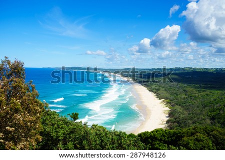 Horizontal view looking onto Tallow Beach in Byron Bay. The Pacific Ocean is a blue turquoise with white waves. The sand is golden, and the beach is surrounded by the green of Arakwai National Park.