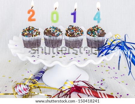 Lit multicolored candles that spell 2014 in chocolate mini cupcakes with colored sprinkles on a cake plate with confetti and party blowouts, noisemakers