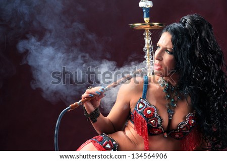 portrait of girl in beautiful clothes east smokes hookah lying on furs on burgundy background studio