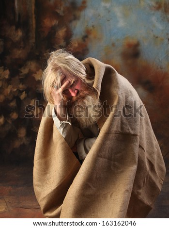 Portrait old beggar with a beard and mustache, wrapped in burlap, with pain and despair in his eyes, the studio on a brown-blue background