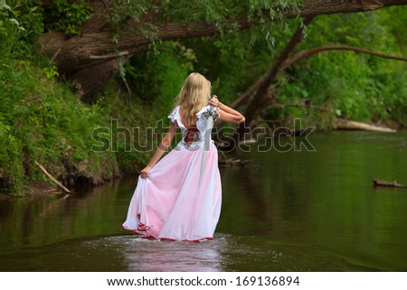 portrait of a beautiful young blonde in a pink dress with a sword in hand, standing in the river in summer day on background green overgrown bushes coast