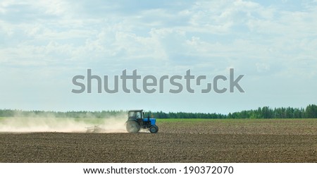 spring landscape blue tractor cultivating a field on a cloudy day and the wood strip on the horizon