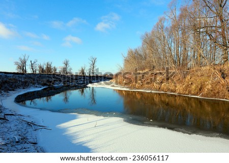 The picturesque landscape of the freezing river and oak trees without leaves on snowy shore on a sunny day in early winter