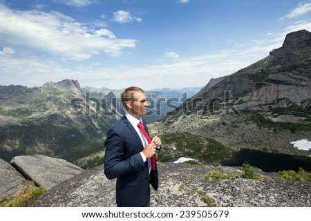 portrait of a young man in a business suit at the top of the mountain on the background of mountains and blue sky on a sunny summer day