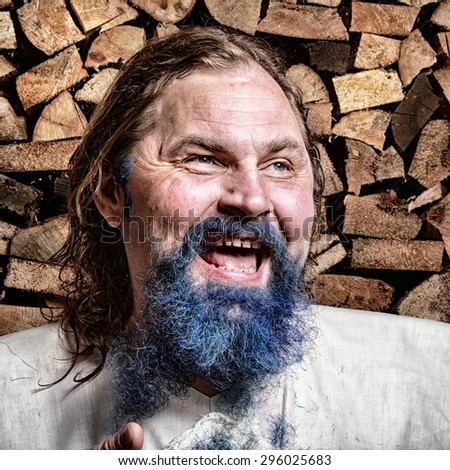 close-up portrait of an adult man with a blue beard and mustache, long blond hair in tissue Cape on background stack of firewood in the bath