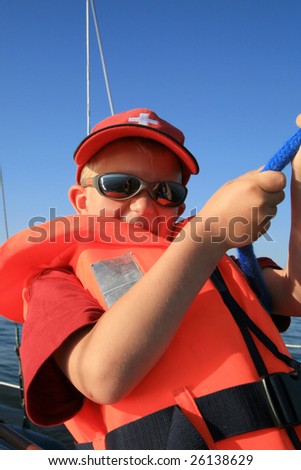 Boy in the orange life jacket with sunglasses on the yacht. He is pulling sheets (ropes). Work aboard, sailing.