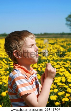 Little boy is blowing dandelion on meadow full of yellow dandelions by may.