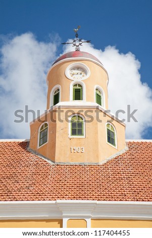 Old clock roof tower with cockerel wind vane on top of red stone roof with blue sky and clouds in background.