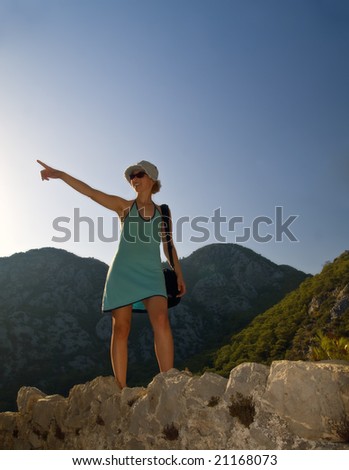 Tourist girl in blue dress walking on rocks in mountains, smiling and pointing on something in front of her