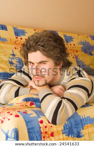 Young smiley dark haired caucasian man in striped sweater lying on country style sofa and frowning