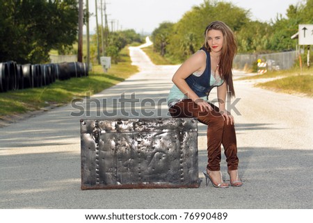 A lovely teenage girl sits fashionably on an antique trunk in the middle of a lonely country road.