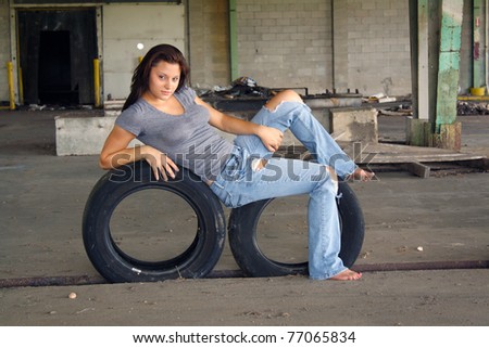 A beautiful young brunette rests on two old tires at an abandoned warehouse facility.
