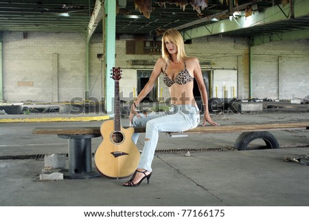 A beautiful blonde in a long-abandoned, dilapidated warehouse sits with her acoustic guitar on a makeshift wooden bench.  She\'s wearing tattered jeans with a leopard-print bikini top.