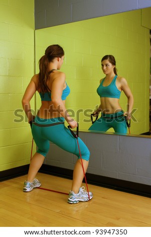 A lovely young brunette with exceptional muscle tone works out with resistance bands in front of a mirror.