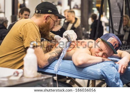 COPENHAGEN, DENMARK - FEBRUARY 8, 2014: a focused tattoo artist tattoos the back of a young man, whose blurred face shakes in pain, in a public location
