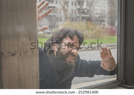 Young handsome bearded man with coat posing behind a glass in the city streets