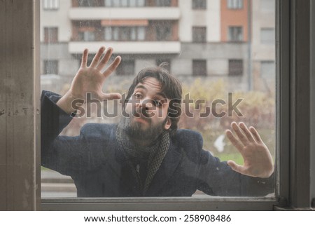 Young handsome bearded man with coat posing behind a glass in the city streets