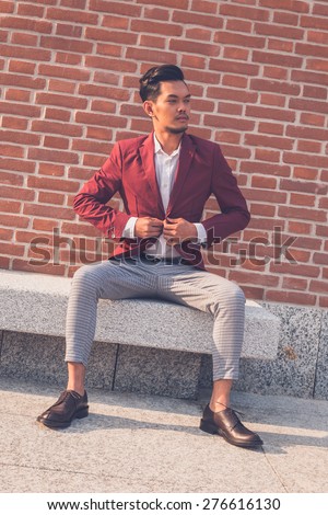 Young handsome Asian model dressed in red blazer sitting on a marble bench in the city streets