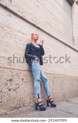 Beautiful blonde girl wearing ripped jeans and leather jacket smoking a cigarette in the city streets