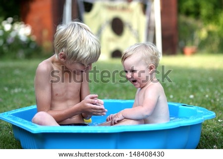 Loving brother with his cute happy baby sister are playing together in a small kiddie pool in the garden, at the backyard of the house