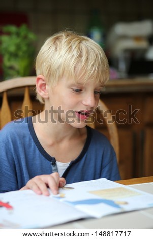 Cute 7 years old boy is studying after school, sitting on a chair in the kitchen and reading his book with home task