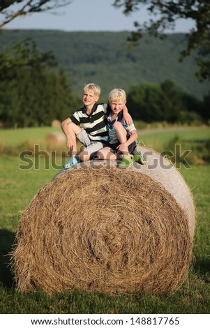 Two cute happy twin brothers are sitting cozy together on top of the hay bale in the middle of the field