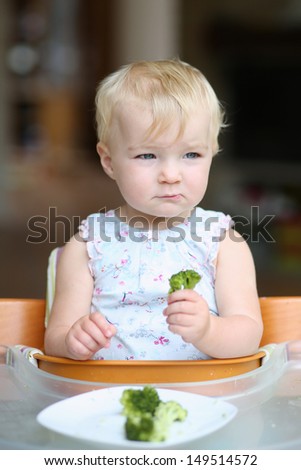 Funny baby girl sitting in a high feeding chair eating steamed broccoli