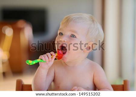 Cute healthy baby with blond hair and blue eyes opens her mouth to eat fresh tomato holding a fork in her hand