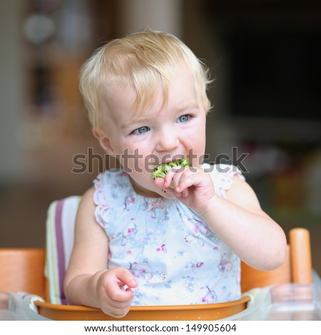 Cute baby girl sitting in a high feeding chair biting on delicious freshly cooked broccoli