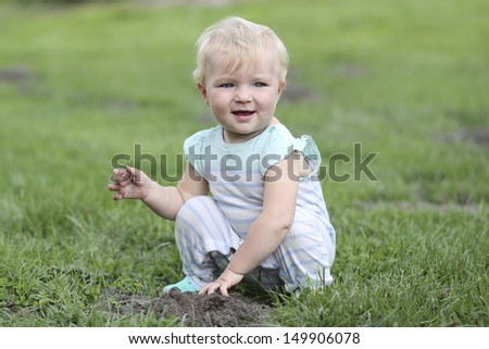 Cute baby girl sitting in a grass in the middle of a big farm playing with mud