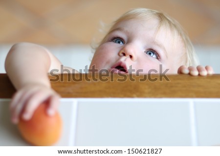 Cute clever baby with beautiful blue eyes and blond hair standing on tiptoes reaching out tasty apricot from high wooden table