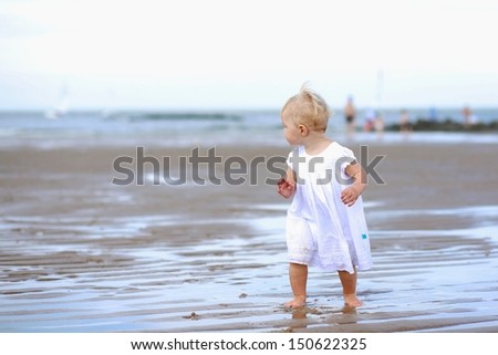 Cute baby girl in white dress running on wet sand along North Sea coastline on a sunny summer day with clear blue sky, she is looking back to the yacht boats in the background, beach Knokke, Belgium