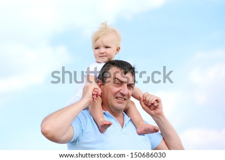 Cute baby girl in white dress sitting on shoulders of happy father, blue sky with white clouds in background