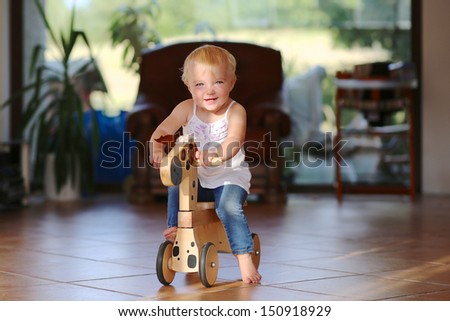 Cute lovely small baby girl learning to balance on her first bike, a wooden horse on wheels. She is standing barefoot on tiles floor inside a house