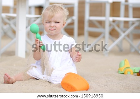 Adorable blond baby girl with white dress plays with toys in the sand at beach cafe