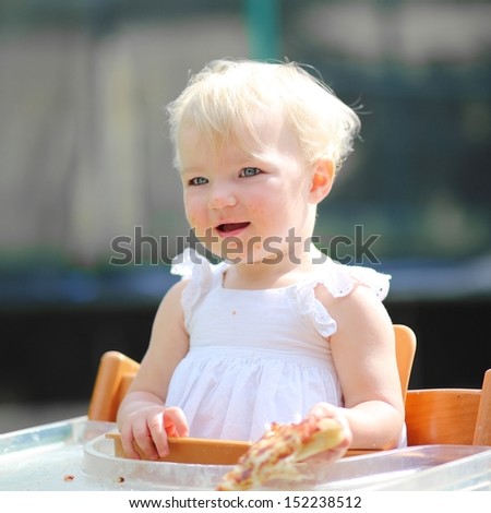Pretty blond little baby girl in beautiful white dress sitting outdoors in feeding chair holding piece of delicious pizza
