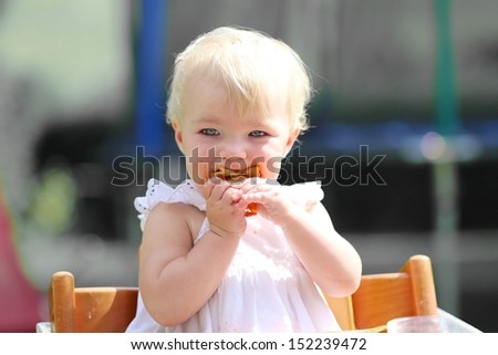 Cute blond little baby girl in beautiful white dress sitting outdoors in feeding chair eating delicious pizza