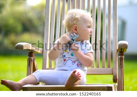 Cute little blond baby girl sitting in a garden on a wooden teak relax chair playing with toy plane