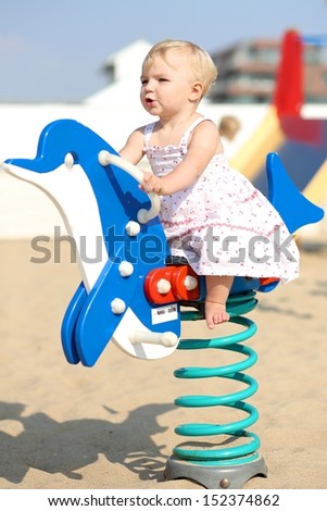 Funny happy blond baby girl plays outdoor at the beach sandy playground rocking on a spring blue dolphin