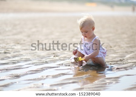 Sweet blond little baby girl in beautiful white dress plays with water and sand sitting at s shore of the sea on a long calm empty peaceful beach, sun is shining at her back