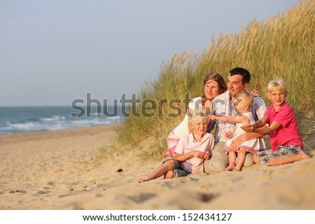 Happy family of five, mother, father, twin brothers and baby daughter are enjoying sunset at a beautiful beach sitting together in dunes