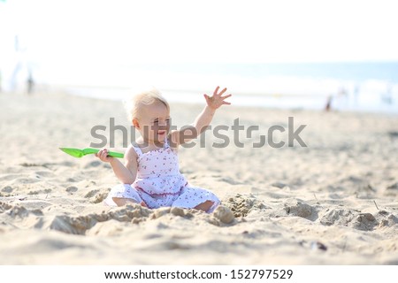 Sweet blond little baby girl in beautiful white dress plays with water and sand sitting at s shore of the sea on a long calm empty peaceful beach