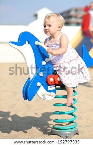 Cute happy blond baby girl plays outdoor at the beach sandy playground rocking on a spring blue dolphin
