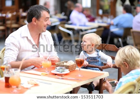 Family of father with children, teenager son and baby daughter are relaxing in cafe on a busy street in the center of the city, laughing and drinking refreshing ice tea and juice