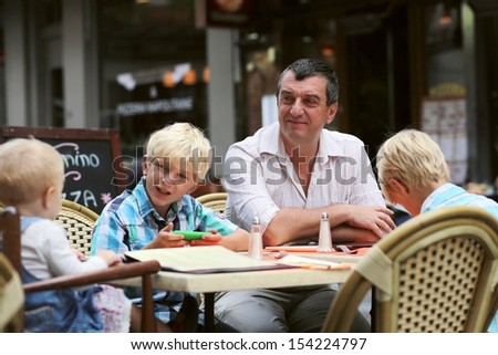 Family, a father with three children, two teenager twin sons and daughter little cute baby girl, are having fun in cafe on summer terrace in a center of busy city street, boy playing with smart phone