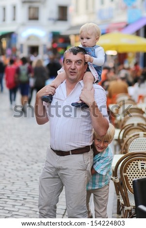 A happy family, father with baby girl daughter on his shoulders and cute teenager boy hiding behind his back are having fun together walking in the middle of the busy city street