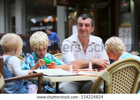 Family, a father with three children, two teenager twin sons and little cute baby girl, are having fun in cafe on summer terrace in a center of busy city street, boy is playing with smart phone
