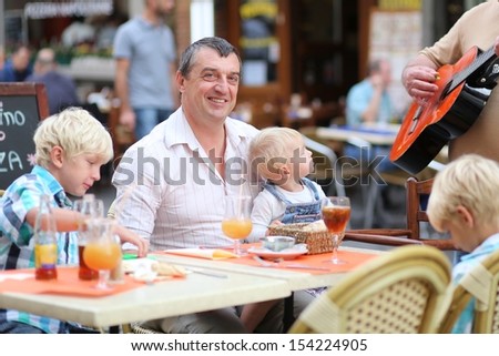 Family, a father with three children, two teenager twins sons and little baby daughter, are listening music in cafe on summer terrace in a center of busy city street, boy is playing with smart phone