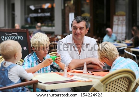 Family, a father with three children, two teenager twins sons, and little cute baby girl, are having fun in cafe on summer terrace in a center of busy city street, boy playing with gadget smart phone