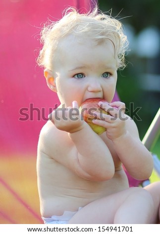 Cute little baby with blond curly hair sitting outdoors in relax chair eating big red apple